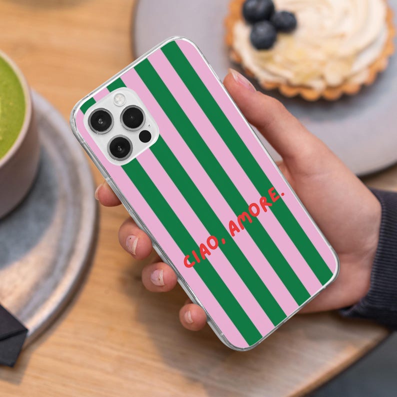 May include: Clear phone case with a pink and green striped pattern. The case has the words "CIAO, AMORE." in red. The phone case is held in a hand.