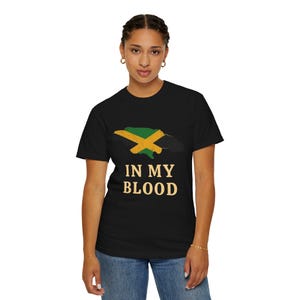 Könnte beinhalten: Schwarzes T-Shirt mit einer grünen und gelben Karte von Jamaika und einem gelben Kreuz. Der Text "IN MY BLOOD" ist unter der Karte in Gold gedruckt. Das T-Shirt wird von einer Person getragen.
