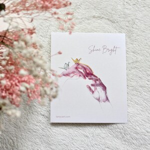 Könnte beinhalten: Ein Aquarelldruck mit zwei Händen, die sich einander entgegenstrecken, mit Origami-Kranichen darauf. Die Worte "Shine Bright" sind kursiv geschrieben. Die Website tanyuart.com ist unten aufgedruckt.