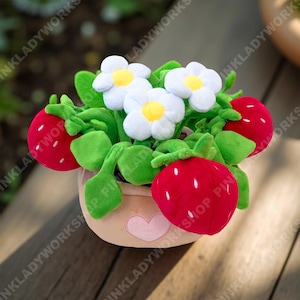 May include: A plush strawberry and flower arrangement in a light brown pot with a pink heart. The strawberries are red with white seeds, and the flowers are white with yellow centers. Green leaves and stems complete the design.