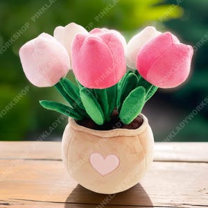 May include: A plush flower arrangement featuring pink and white tulip blossoms with green leaves, set in a beige pot with a heart applique. The soft toy is designed to resemble a potted plant.