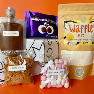 May include: A selection of ingredients for making waffles, including a pouch of waffle mix, chocolate sauce, Biscoff crumb, marshmallows, and a Cadbury Dairy Milk Buttons chocolate bar. The waffle mix package has the text "Waffle Mix".