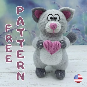 以下が含まれることがあります： ピンクのハートを持った、かぎ針編みの灰色の猫。猫は、ピンクの内側の耳、ピンクの鼻、大きな目を持つ灰色の耳を持っています。「FREE PATTERN」という言葉が、左側に紫色の文字で縦に書かれています。