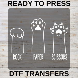May include: White DTF transfers with a gray wood grain background. The design features three hand-drawn paw shapes representing Rock, Paper, and Scissors, with the words below each shape. The text "READY TO PRESS" and "DTF TRANSFERS" are also included.
