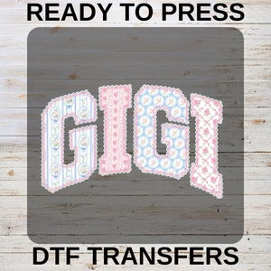 May include: DTF transfer featuring the name "GIGI" in a decorative font. The letters are filled with floral and patterned designs in pink and blue, with a scalloped edge. The words "READY TO PRESS" and "DTF TRANSFERS" are at the top and bottom.