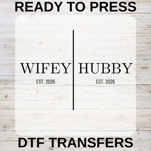 May include: White DTF transfer featuring the words "Wifey" and "Hubby" divided by a vertical line. The text "Est. 2026" is below each word. The phrases "Ready to Press" and "DTF Transfers" are also displayed.