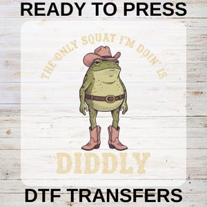 May include: A DTF transfer design featuring a cartoon frog wearing a cowboy hat, boots, and a belt. The text reads "The only squat I'm doin' is diddly". The words "READY TO PRESS" and "DTF TRANSFERS" are also included.