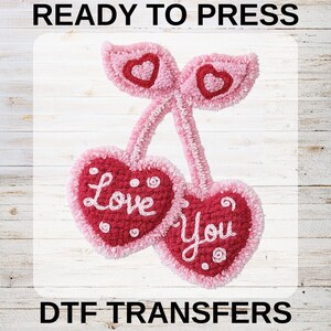 May include: A pink and red heart-shaped DTF transfer with the words "Love You" in white script. The transfer features a cherry design with a heart-shaped leaf and a heart in the center of each cherry. The words "READY TO PRESS" and "DTF TRANSFERS" are printed above and below the design.
