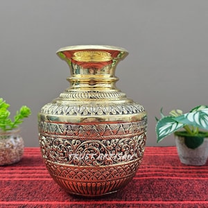 May include: A gold-colored, ornate vase with intricate floral patterns. The vase has a wide base, a rounded body, and a flared rim. The vase is sitting on a red striped surface.
