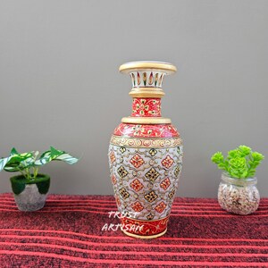 Può includere: Un vaso decorativo dipinto a mano con una parte superiore e inferiore color crema, una sezione centrale rossa e dorata e un corpo bianco ornato da motivi floreali. Il vaso ha il testo "TRUST ARTISAN" sul davanti.