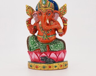 May include: A hand-painted orange Ganesha figurine featuring a green patterned dhoti, seated on a pink lotus base with a small black mouse at the front. The deity wears a golden crown and jewelry, holding a golden staff and a wheel-shaped emblem.