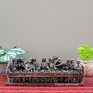 May include: A bronze-colored sculpture depicting the Last Supper. The detailed figures are seated around a long table, with the text "TRUST ARTISAN" at the base. The sculpture is set against a neutral background with decorative plants.