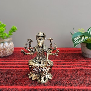 May include: A detailed bronze-colored statue of a deity with multiple arms, seated on a lotus flower base. The statue has intricate detailing and is adorned with a headdress and jewelry. The word "ARTISAN" is visible on the base. The statue is on a red striped surface.