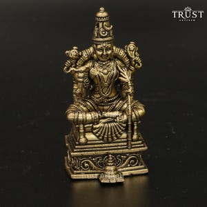 May include: A metallic bronze-toned figurine of a seated deity with four arms, wearing an ornate crown and jewelry. The figure sits on a rectangular pedestal with intricate carvings. The text TRUST ARTISAN is visible in the top right corner.