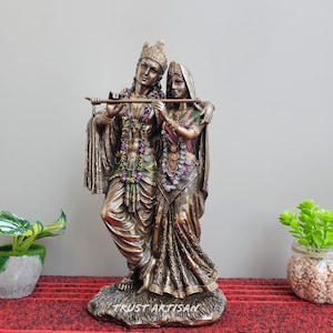 May include: A bronze-colored statue of a couple, likely representing a deity, stands on a textured base. The male figure holds a flute, while the female figure is draped in a sari. The statue is adorned with intricate details and jewelry. The base has the text "TRUST ARTISAN".