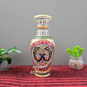 May include: A decorative, hand-painted marble vase with a cream-colored base and rim. The vase features a detailed design of two peacocks surrounded by floral patterns in red, green, blue, and gold. The vase is approximately 12 inches tall.