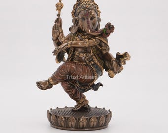 May include: A bronze-colored statue of the deity Ganesha in a dancing pose, standing on a lotus-shaped base. The figure features intricate clothing details, jewelry, and holds a mace in one hand, with the text Trust Artisan visible.