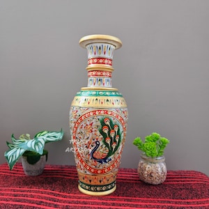 May include: A tall, decorative vase with a cream-colored base and top. The vase features a detailed peacock design in red, green, and blue, with intricate patterns. The vase is placed on a red striped surface.