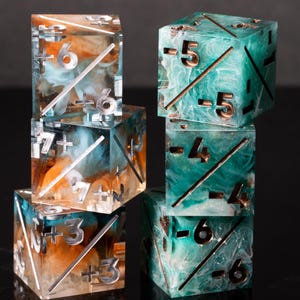 May include: A set of six translucent resin dice stacked in two columns. The left column features orange and white swirls with silver-colored positive numbers and diagonal lines. The right column features teal and white swirls with copper-colored negative numbers.