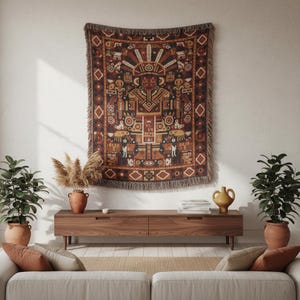 May include: A woven tapestry with a geometric and figurative design in shades of brown, orange, and black hangs on a white wall. The tapestry has a fringed edge. The room features a wooden cabinet, plants, and a sofa.