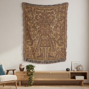 May include: A woven tapestry with a detailed, brown and tan geometric design featuring a central figure with elaborate headwear and holding objects. The tapestry has a fringed edge and is displayed on a white wall. The overall theme is ethnic and decorative.