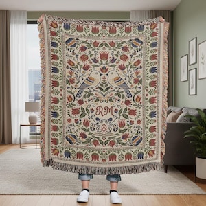 May include: A woven throw blanket with a floral and bird design. The blanket features a cream background with red, blue, and green floral patterns. Two birds are centered above a heart-shaped design with the initials "RM". The blanket has a fringed edge.