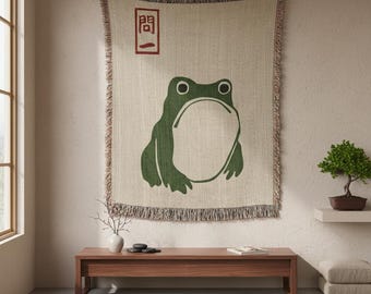 May include: A beige throw blanket with a large green frog illustration. The blanket has a fringed edge and a red Japanese character in the upper left corner. The frog has a large open mouth. The blanket is hanging on a wall.