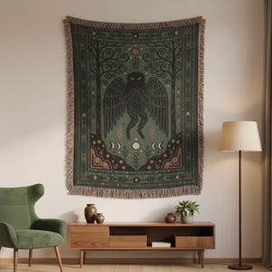 May include: A woven tapestry featuring a dark green background with a central illustration of a winged creature. The design includes trees, moons, and floral elements in shades of black, brown, and cream. The tapestry has a fringed edge.