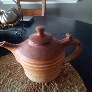May include: A brown ceramic teapot with a lid and a curved handle. The teapot has a speckled design and is sitting on a woven placemat. The teapot is a warm, earthy color.