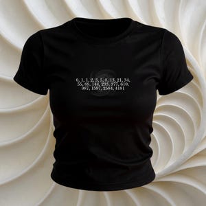 Puede incluir: Camiseta negra de cuello redondo con texto blanco y un diseño circular. El texto incluye una serie de números: 0, 1, 1, 2, 3, 5, 8, 13, 21, 34, 55, 89, 144, 233, 377, 610, 987, 1597, 2584, 4181. La camiseta es de material suave.