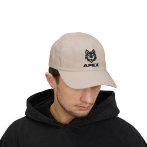 May include: A beige baseball cap featuring a black graphic of a wolf head centered above the word APEX in bold, black, sans-serif capital letters. The cap has a structured crown, a curved brim, and a button at the top center of the fabric dome.