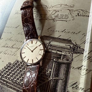 May include: A classic wristwatch featuring a silver-toned face and a brown leather strap. The watch has a gold-coloured buckle and is set against a backdrop of a postcard and typewriter illustration. The watch face displays the brand name "UNIVERSAL GENEVE".