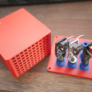 Könnte beinhalten: Eine rote Audiokomponente mit quadratischem, belüftetem Design, neben einer rot-blauen Basis mit drei Audiokartuschen. Die Komponente trägt das Wort "THORENS" in goldfarbenen Buchstaben. Die Patronen haben silberne und schwarze Details.