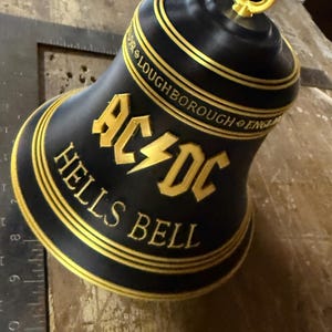 May include: A black bell with gold trim and the text "AC/DC HELLS BELL" in gold lettering. The bell has a gold ring at the top and the text "LOUGHBOROUGH ENG" around the top rim.