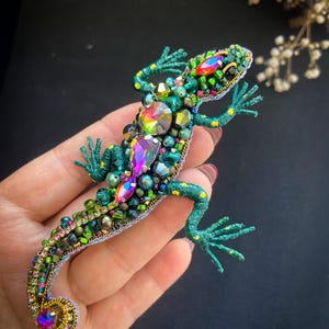 Puede incluir: Broche de lagarto hecho a mano con cuentas en tonos verdes, turquesas y dorados. El lagarto está adornado con cristales y cuentas iridiscentes y coloridas. Un accesorio único y llamativo.