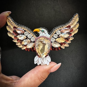 Puede incluir: Un broche de águila detallado con las alas extendidas. El broche presenta una gran gema amarilla en forma de lágrima en el centro, rodeada de piedras más pequeñas transparentes y color ámbar. Las alas están adornadas con lentejuelas doradas y rojas, y la cabeza del águila es blanca con un pico amarillo.