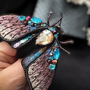 Puede incluir: Broche de polilla artesanal con detalles intrincados. Las alas son una mezcla de marrón y morado, adornadas con lentejuelas y abalorios. El cuerpo es negro, adornado con una gran gema en forma de lágrima y piedras azules y rosas más pequeñas.