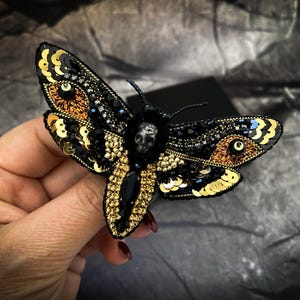 Puede incluir: Broche de polilla artesanal con detalles intrincados. El accesorio en forma de insecto, negro y dorado, presenta lentejuelas, cuentas y un acento de calavera. Las alas tienen un patrón dorado y negro, y el cuerpo tiene una gema negra.