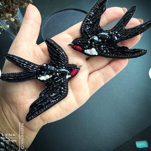 Puede incluir: Dos broches de golondrina negros con cuentas y detalles intrincados. Las aves tienen acentos rojos en sus picos y blancos en sus pechos. Los broches están adornados con cuentas y lentejuelas negras, creando un efecto texturizado y brillante. Los broches están hechos a mano.