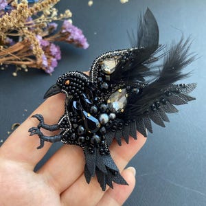 Puede incluir: Broche de cuervo negro, intrincadamente adornado con cuentas negras, cristales y plumas. Las alas del ave están extendidas y su cuerpo está adornado con cuentas y cristales negros de varios tamaños. Un accesorio único.