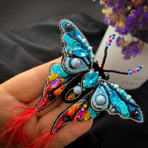 Puede incluir: Broche de mariposa artesanal con intrincado trabajo de cuentas en tonos azules, turquesas e iridiscentes. Las alas presentan lentejuelas y cristales, con detalles de plumas rojas. Un accesorio único.