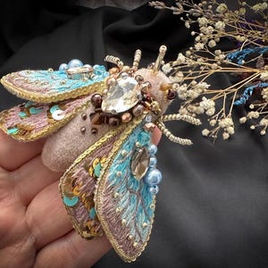 Puede incluir: Broche artesanal con forma de polilla, con intrincados bordados en tonos azules, rosas y dorados. El broche está adornado con cuentas y un gran cristal transparente. Las alas tienen un aspecto detallado y texturizado.
