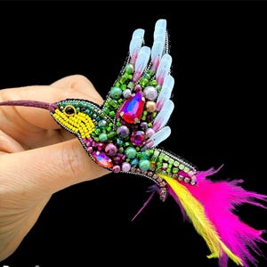 Puede incluir: Broche de colibrí hecho a mano con un diseño vibrante. El broche presenta un cuerpo de cuentas amarillas, verdes y moradas, un pico rojo y una cola de plumas rosas y amarillas. Las alas están adornadas con detalles blancos y transparentes. Un accesorio único.
