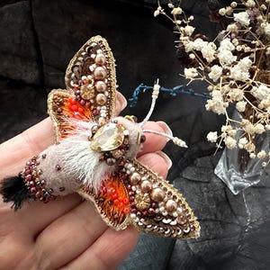Puede incluir: Broche de polilla hecho a mano con intrincados abalorios en tonos marrón, naranja y dorado. La polilla presenta una gran gema transparente y detalles de piel blanca. Un accesorio artístico y único para ropa.