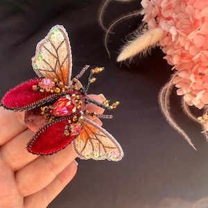 Puede incluir: Broche de insecto bordado a mano, con alas de terciopelo rojo y detalles de cuentas. Las alas presentan intrincadas costuras en tonos naranja, beige y verde. El broche está adornado con cuentas rosas y doradas, creando un accesorio único.