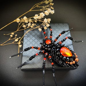 Puede incluir: Broche de araña con cuentas con cuerpo negro, detalles rojos y detalles de piedras preciosas naranjas. La araña está sobre una superficie negra texturizada, con una ramita de flores secas en el fondo. Un accesorio llamativo.