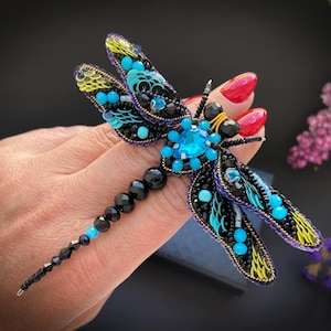 Puede incluir: Broche de libélula con cuentas detalladas. Las alas están adornadas con cuentas turquesas, negras y amarillas, mientras que el cuerpo presenta cuentas negras y azules. Un gran cristal azul claro se encuentra en el centro.