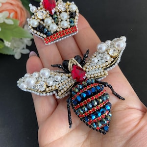 Puede incluir: Un broche de abeja enjoyado y un broche de corona. La abeja tiene alas de perlas, una gema roja y un cuerpo enjoyado azul y rojo. La corona está adornada con perlas, gemas rojas y cristales transparentes.