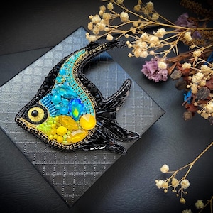 Puede incluir: Broche artesanal con forma de pez, con un contorno de cuentas negras. El pez presenta un diseño de cuentas turquesas, amarillas y verdes, con un borde de cuentas doradas. El broche está sobre una superficie gris texturizada, con flores secas en el fondo.