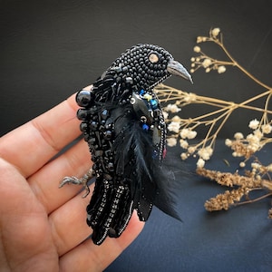 Puede incluir: Broche artesanal de cuervo negro con cuentas y diseño detallado. El ave presenta un cuerpo texturizado, un ojo prominente y detalles de plumas y cuentas de colores. Un accesorio único.